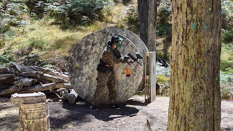 A person in tactical gear hiding behind a wooden barrel at a paintball field.