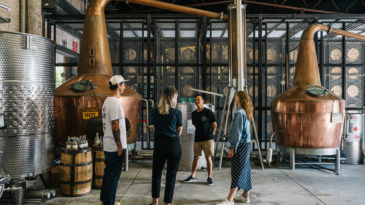 A group of people stand in a distillery surrounded by two large copper stills.