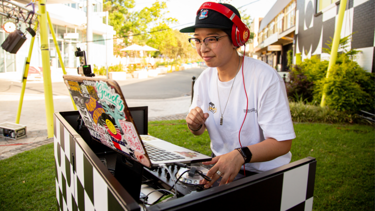 A woman wearing a white t-shirt, glasses, a black hat and red headphones DJs on a laptop.