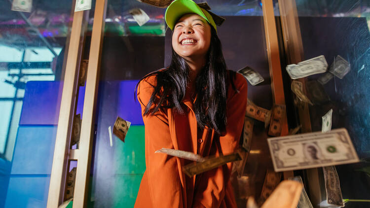 A girl wearing a green cap and orange jacket standing in a room with dollar bills falling. 