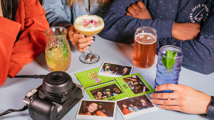 A group of friends sitting around a table with cocktails and polaroid pictures.