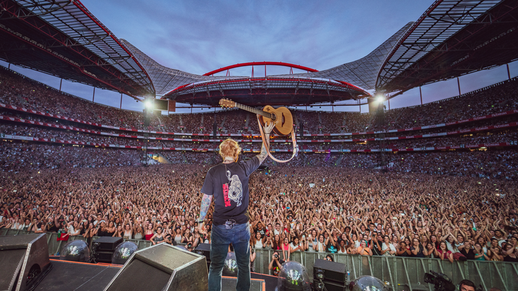 Ed Sheeran holding a guitar on stage in front of a huge crowd.