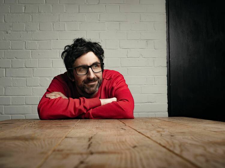 Coemdian Mark Watson leaning on a table while wearing a red sweatshirt.