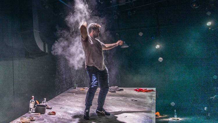 A man surrounded by a smoky haze stands on a stage littered with empty bottles, rubbish and clothes.