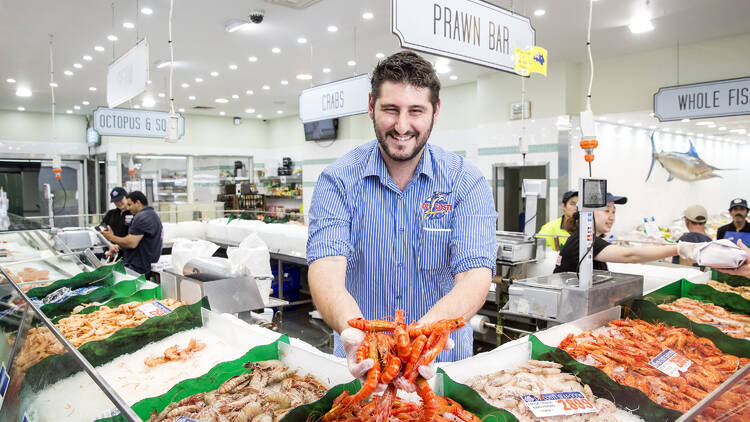 A smiling man holds out a handful of prawns to the camera in a brightly lit seafood shop
