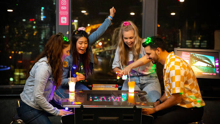 A group of three woman and one man wearing '80s-inspired clothing play an old-school arcade game.