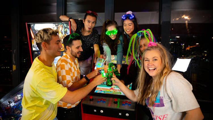 A group of people dressed in '80s gear cheers their drinks over an old-school arcade game.
