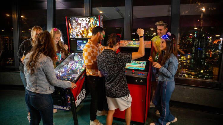 A group of people dressed in '80s gear play on some old-school arcade games.