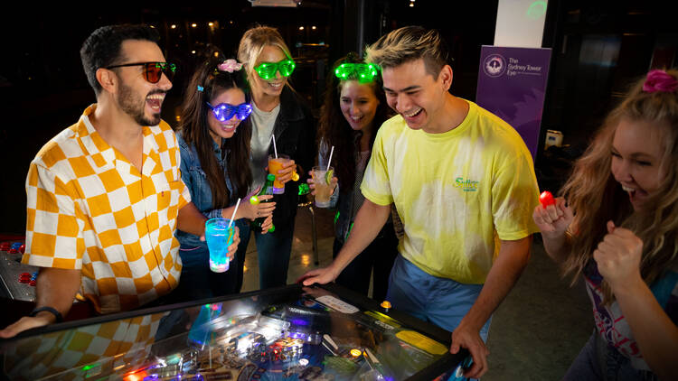 A group of people dressed in '80s gear watch on as a man in a yellow t-shirt plays an old-school pinball machine.