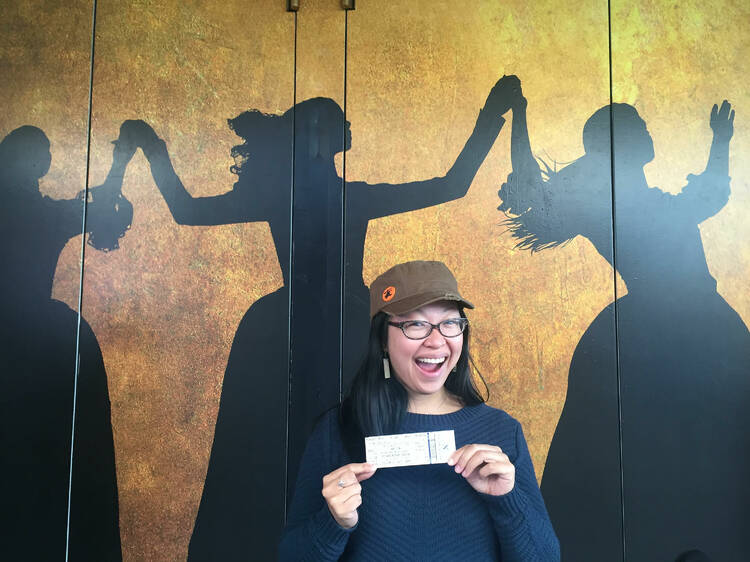 A woman with a big smile holding a Hamilton ticket in front of the Hamilton artwork