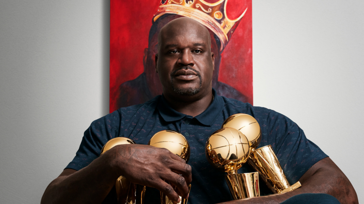 Shaquille O'Neal sits in front of a portrait, only a tilted crown visible while he holds his trophies