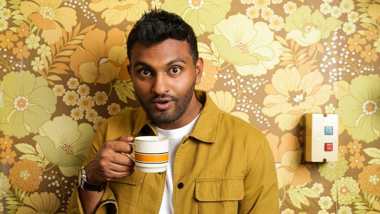 Nazeem Hussain stands in front of a yellow floral wall with a cup of tea