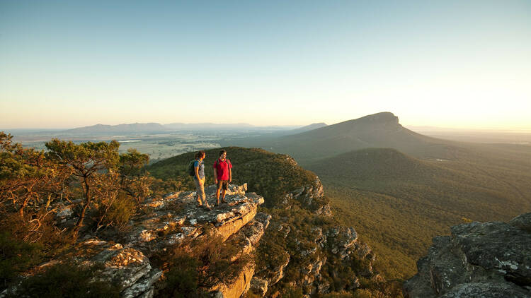 Two hikers stand on top of a rocky cliff, with dramatic mountain peaks in the background.