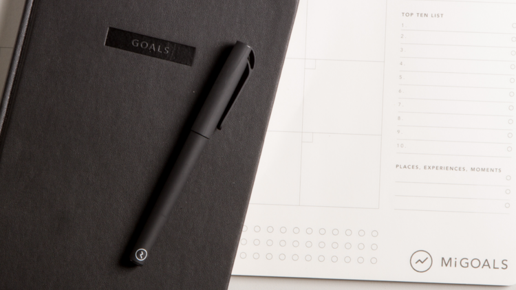 A black journal with a black pen sits on top of a large daily planner.