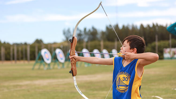 A boy in a blue and yellow singlet practices archery on a green field.