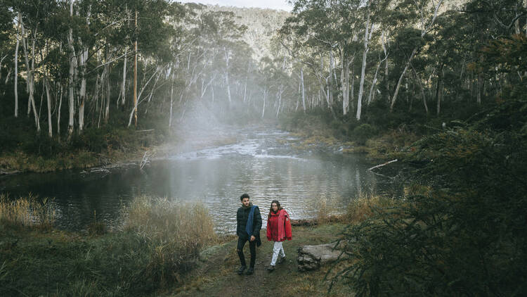 A man in a dark green jacket and a woman in a red jacket walk past a misty lake in the bush.