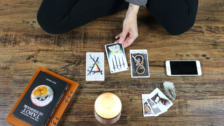 Woman sitting on wooden floor with tarot card spread, reading her fortune surrounded by candle burning, phone, photos and notebook. Eastern philosophy, alternative worship at an altar of sorts.