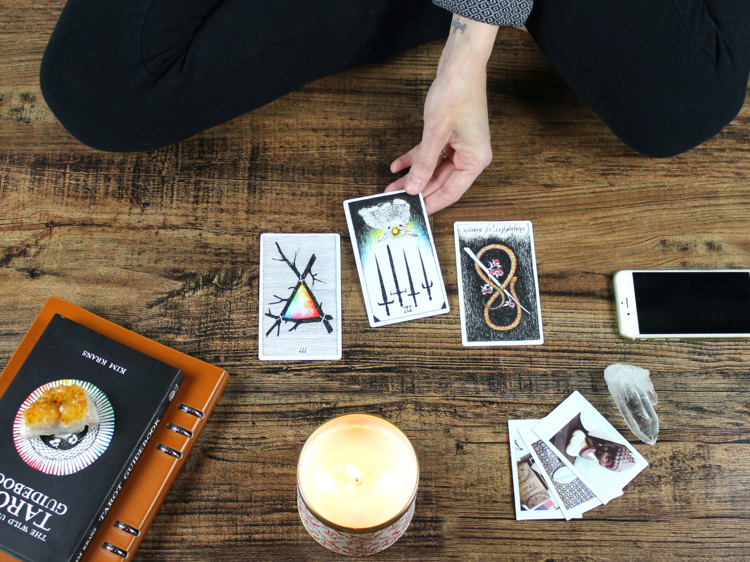 Woman sitting on wooden floor with tarot card spread, reading her fortune surrounded by candle burning, phone, photos and notebook. Eastern philosophy, alternative worship at an altar of sorts.