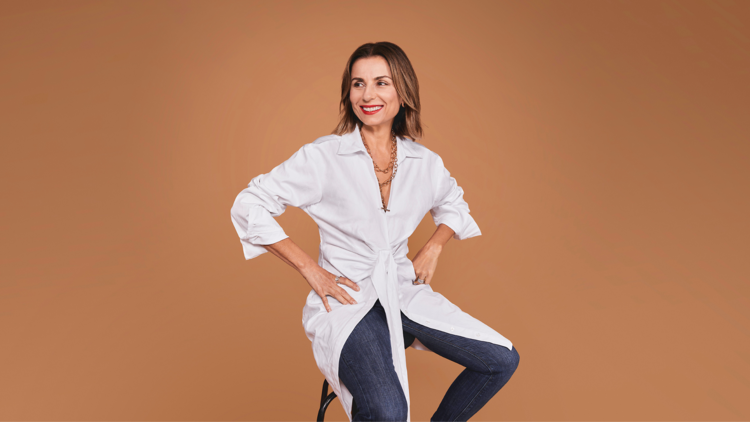 A woman with shoulder-length brown hair and wearing a white shirt sits on a stool against a light-brown backdrop.