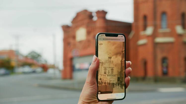 A person holding a phone up with a historic photo on it, against a historic building.