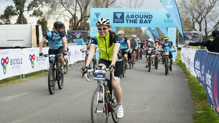 A smiling woman on a bike pedals out in front of a larger group of riders.