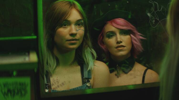A still from Poser Still; two women staring at their reflections in a mirrored surface.
