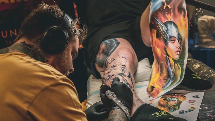 A man wearing headphones tattoos a person's leg.