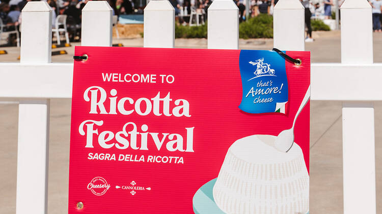 A white fence with a red sign that reads 'welcome to Ricotta Festival'.