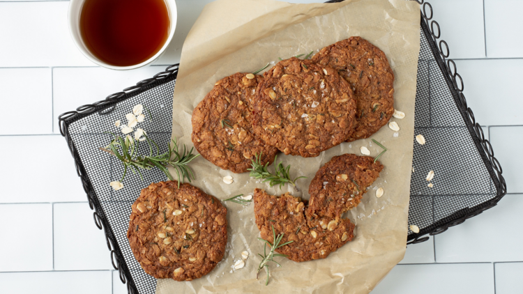 A cooling rack with anzac biscuits and rosemary next to a cup of tea