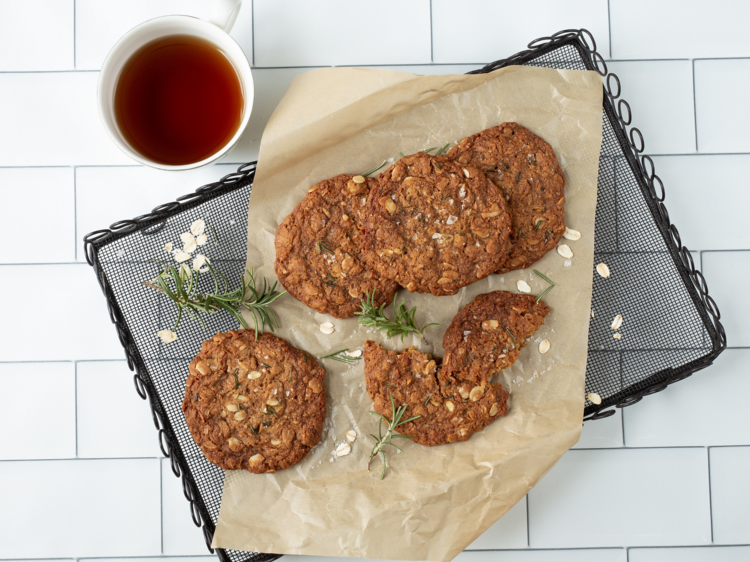 A cooling rack with anzac biscuits and rosemary next to a cup of tea