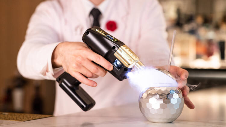 A bartender in a white suit holds a smoke machine up to a disco ball-shaped cup.