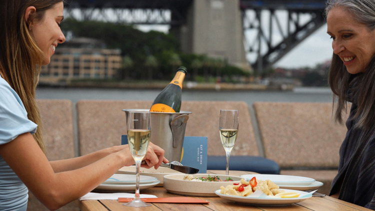 A mother and daughter sit at Opera Bar, the Harbour Bridge in the background, drinking Champagne