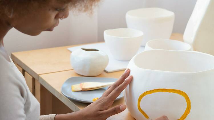 A woman paints a yellow circle on a ceramic pot. More unpainted pots are resting on the table in the background.