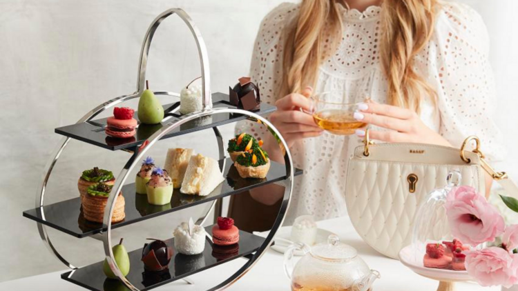 A woman holding a cup of tea sits next to a three-tiered tray filled with scones, sandwiches and other sweet treats.