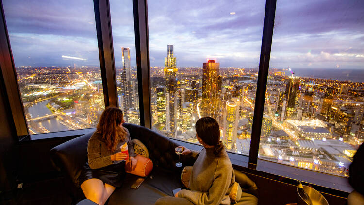 Two women sitting on a couch looking out the window at a view of a city skyline.