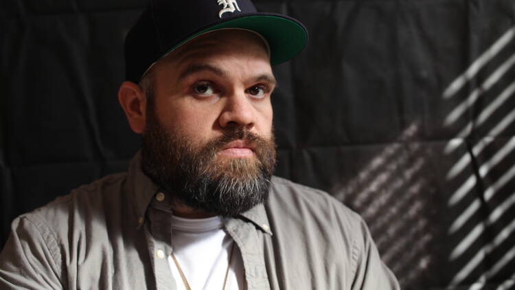 Rapper Briggs wears a baseball cap, grey shirt on a dark backdrop