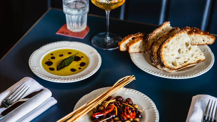 A dining table loaded with plates of bread, olives, dipping oil, cutlery and glasses of wine.