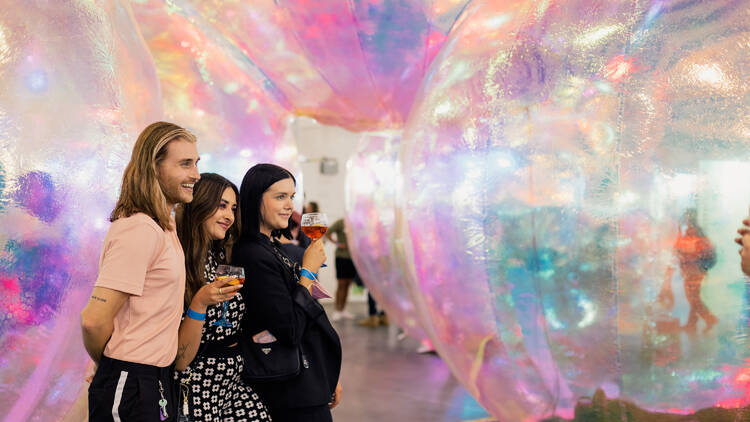 Three people holding drinks pose together among giant translucent bubbles.