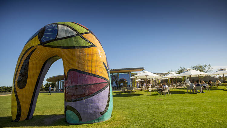 A colourful, semi circular sculpture sits in the foreground on grass, with people sitting under umbrellas in the background