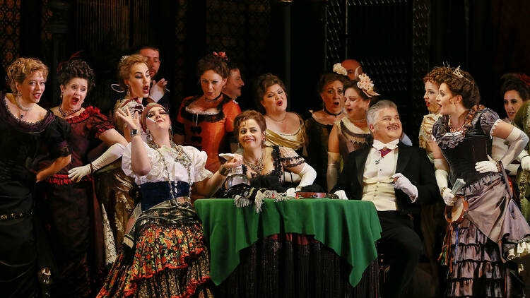 The cast of La Traviata sit around a table with a green tablecloth, animated and interacting