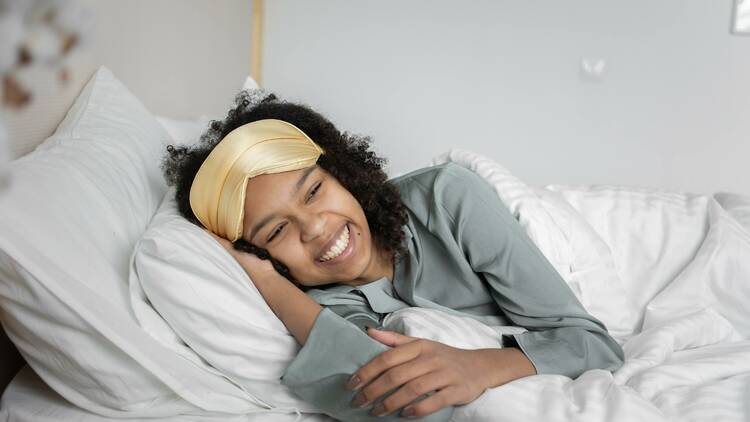 A woman laying in bed with white linens while wearing a green pyjama top and yellow eye mask.