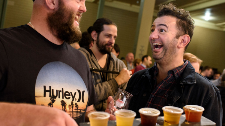 Two men laugh at each other over a tray of a beer tasters 