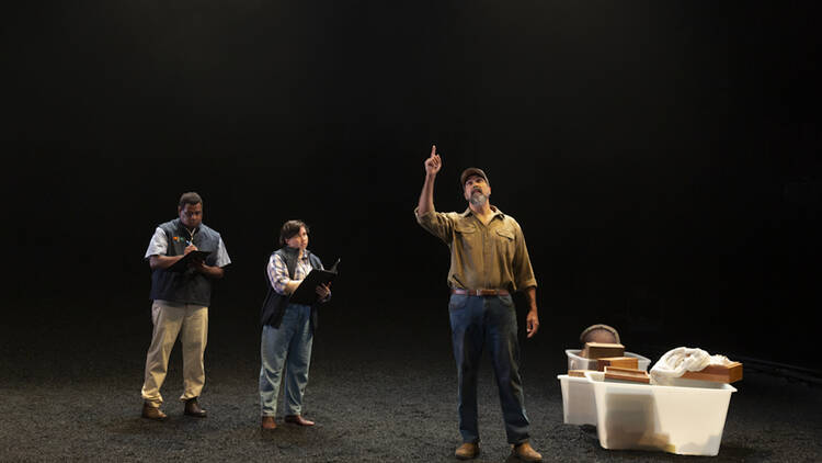 A man stands in darkness, pointing to the sky, as two people with clipboards write something down