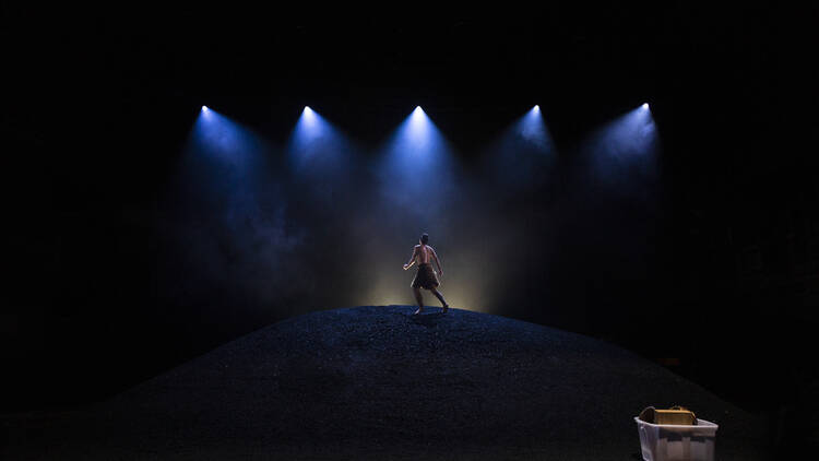 A man in a fur wrap skirt stands in the darkness atop a large mound, spotlights shining down on him, his back to the audience