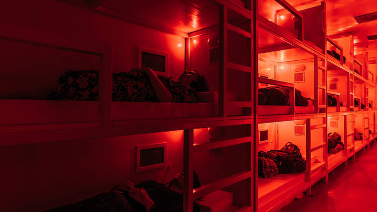 A shipping container is filled with bunk beds. People lay in the beds bathed in red light.