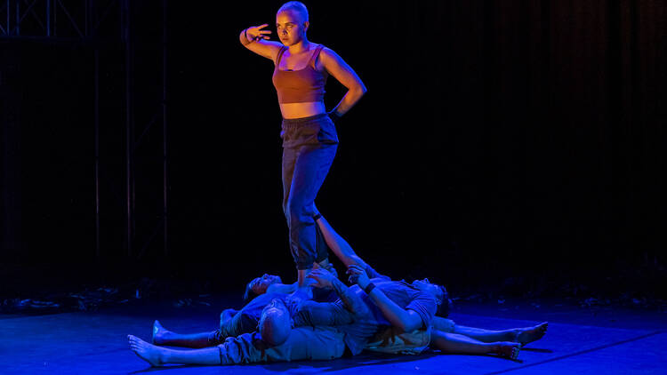 A woman stands, looking towards a spotlight, being held up by a group of dancers collapsed on the floor