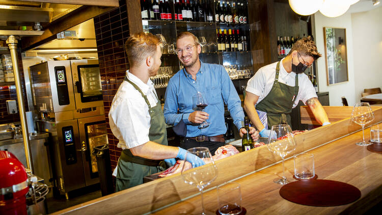 Two men in aprons and one in a blue shirt stand at a bar holding glasses of wine.