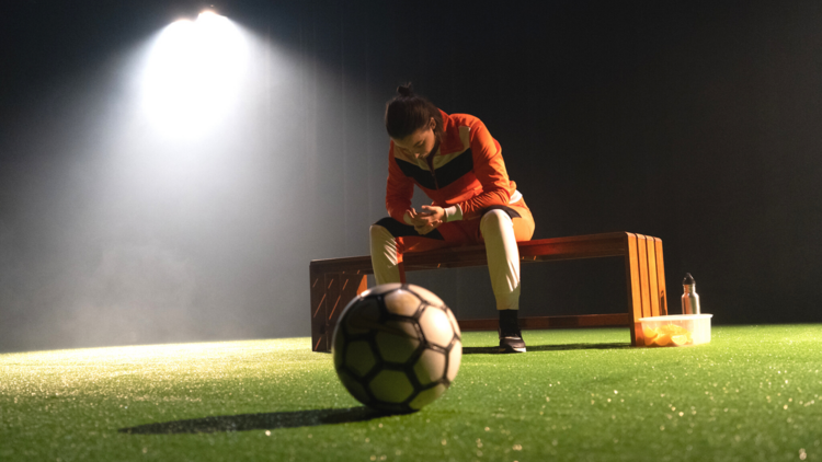 Jamila Mainwears an orange tracksuit and sits on a park bench on an astro turf field, a soccer ball is in the foreground