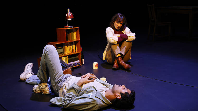 A man lies on the floor, next to a bookcase, and a women looks down at him, sitting crosslegged