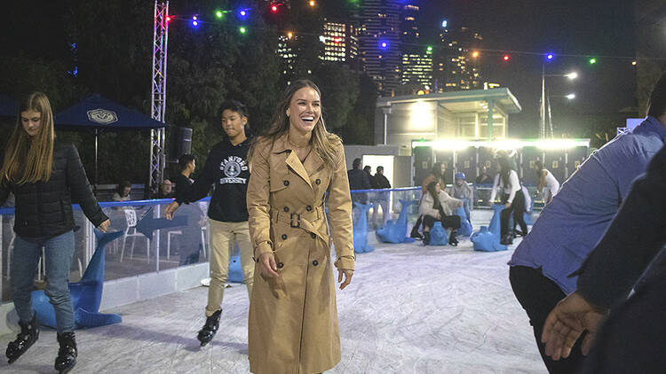 A woman in a camel trench coat ice-skating.